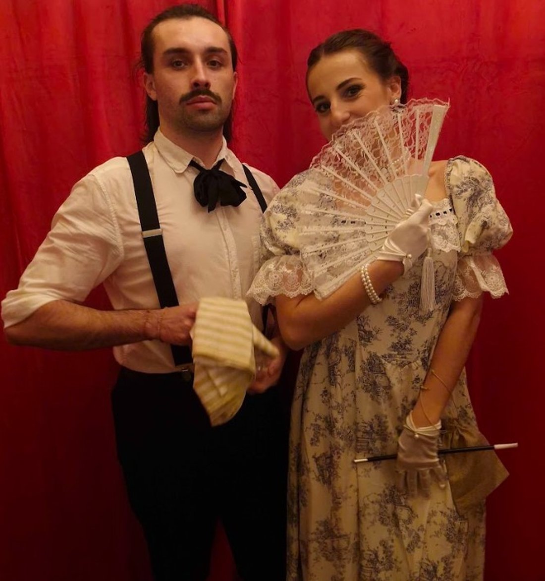 Deux comédiens costumés en gentleman et débutante Belle Époque devant un rideau rouge