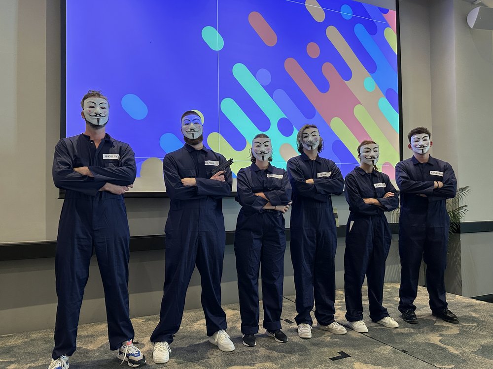 Équipe de six joueurs masqués en combinaison face à un écran de données, prête à infiltrer le réseau
